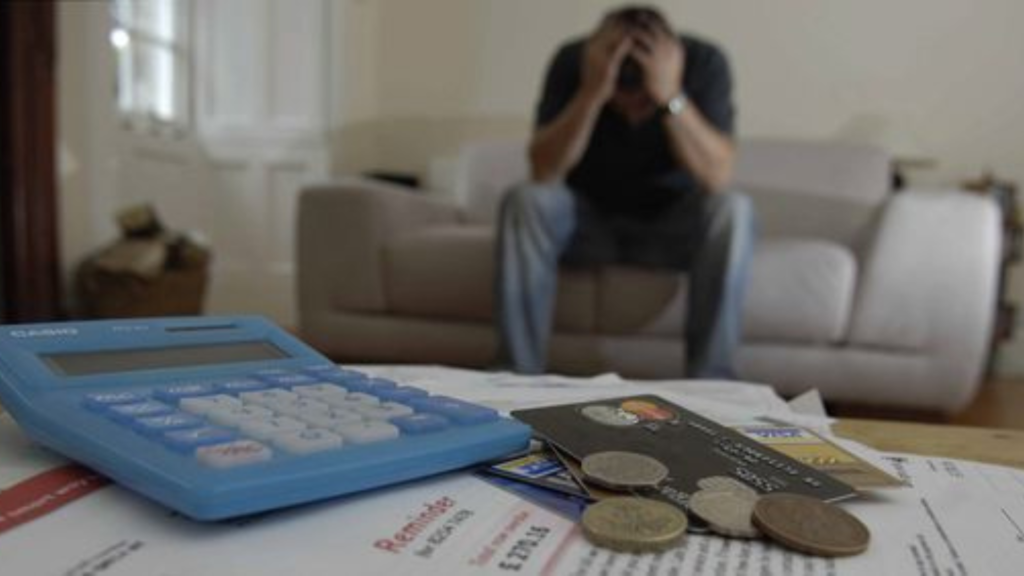 A frustrated man sits on a chair with calculator, documents and coins on the table