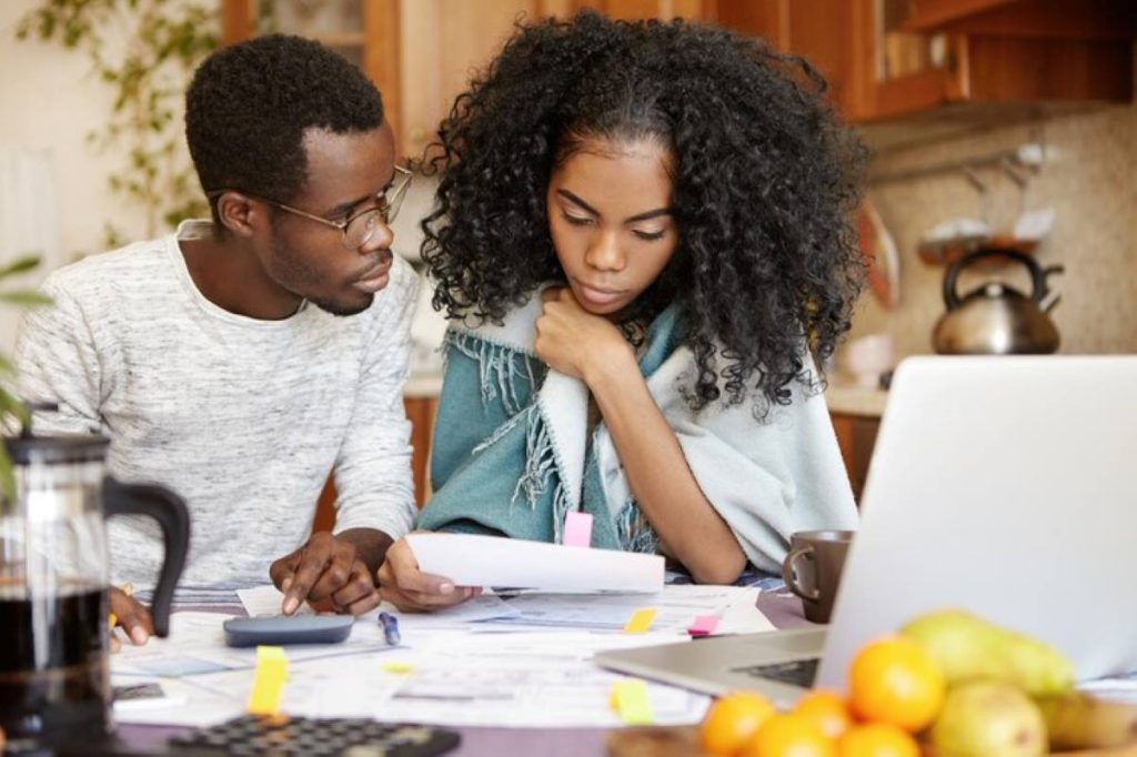 Dark-skinned male holding pencil, making calculations on a calculator and looking at his worried wife